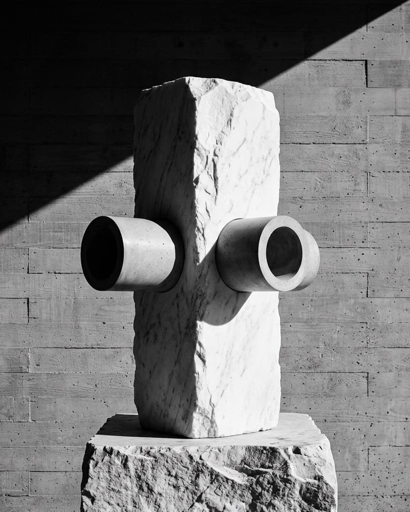 Un bloc de marbre blanc traversé de part en part par deux tubes de béton, posé sur un socle de pierre, devant un mur de béton banché.