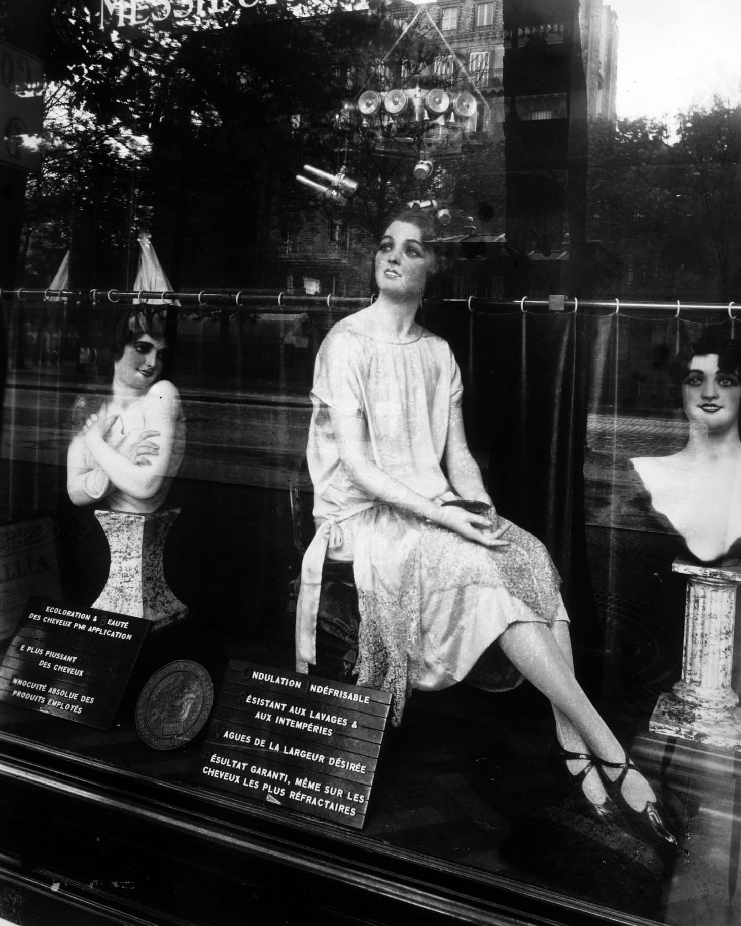 La vitrine d’un coiffeur parisien : des bustes et un mannequin assis présentent les coiffures, derrière des affichettes.