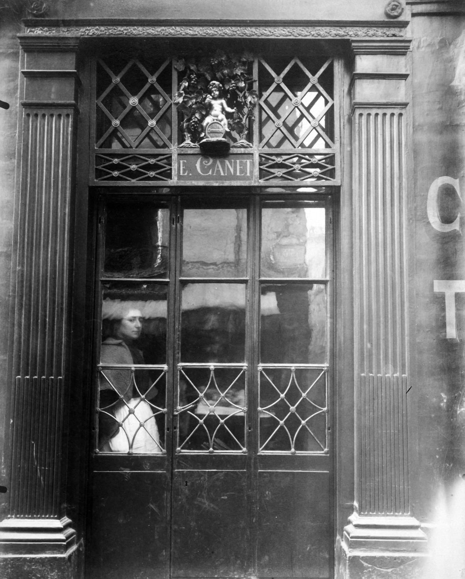 La devanture d’une boutique parisienne : une porte vitrée sous une grille de fer forgé, une enseigne sculptée au-dessus.