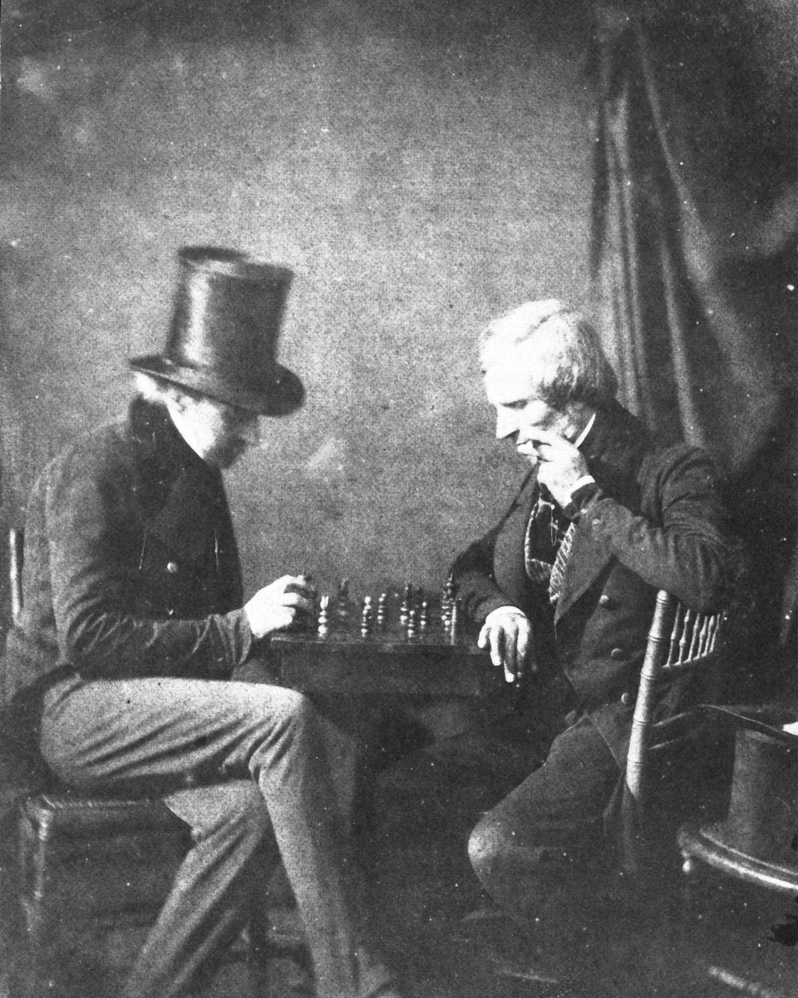 Deux hommes en redingote penchés sur un échiquier, l’un coiffé d’un haut-de-forme, l’autre la main au menton, devant une draperie.