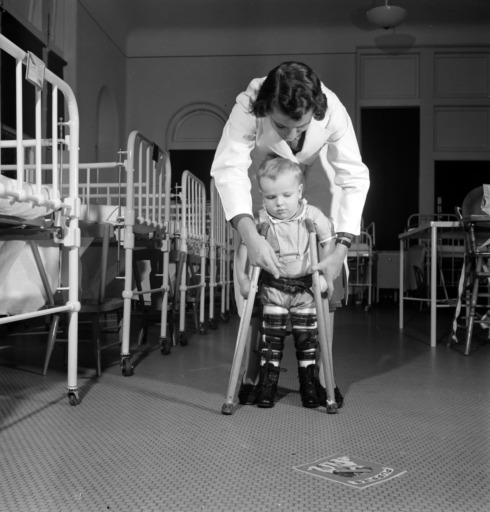 Une soignante guide un enfant en appareil orthopédique qui apprend à marcher avec des béquilles, dans une salle d’hôpital.