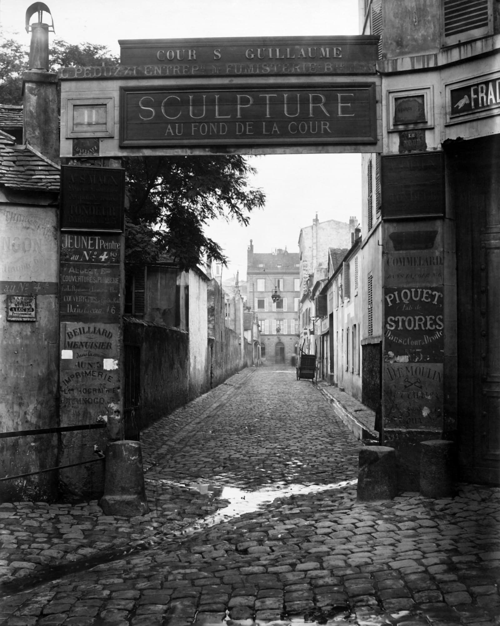 Un porche pavé ouvrant sur une ruelle ; au-dessus, une grande enseigne « Sculpture au fond de la cour » et, sur les murs, les plaques des artisans installés derrière.