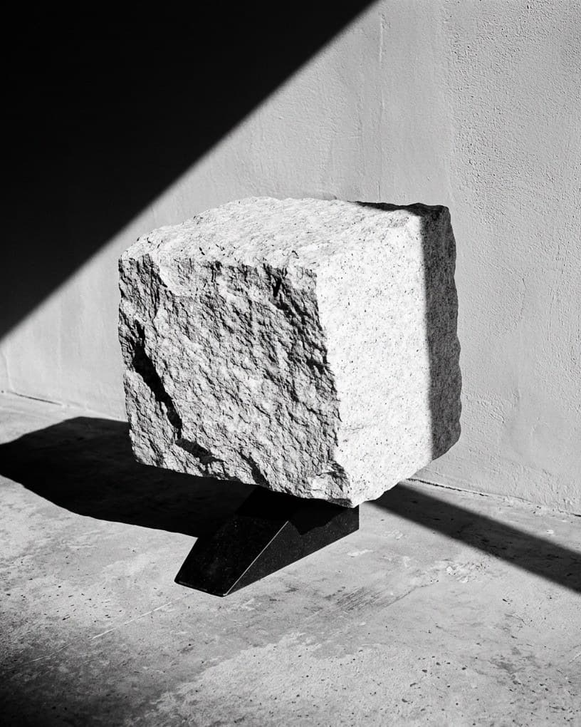 Un bloc de granit taillé en équilibre sur une cale de basalte noir, au pied d’un mur blanc, une ombre en diagonale sur le sol.