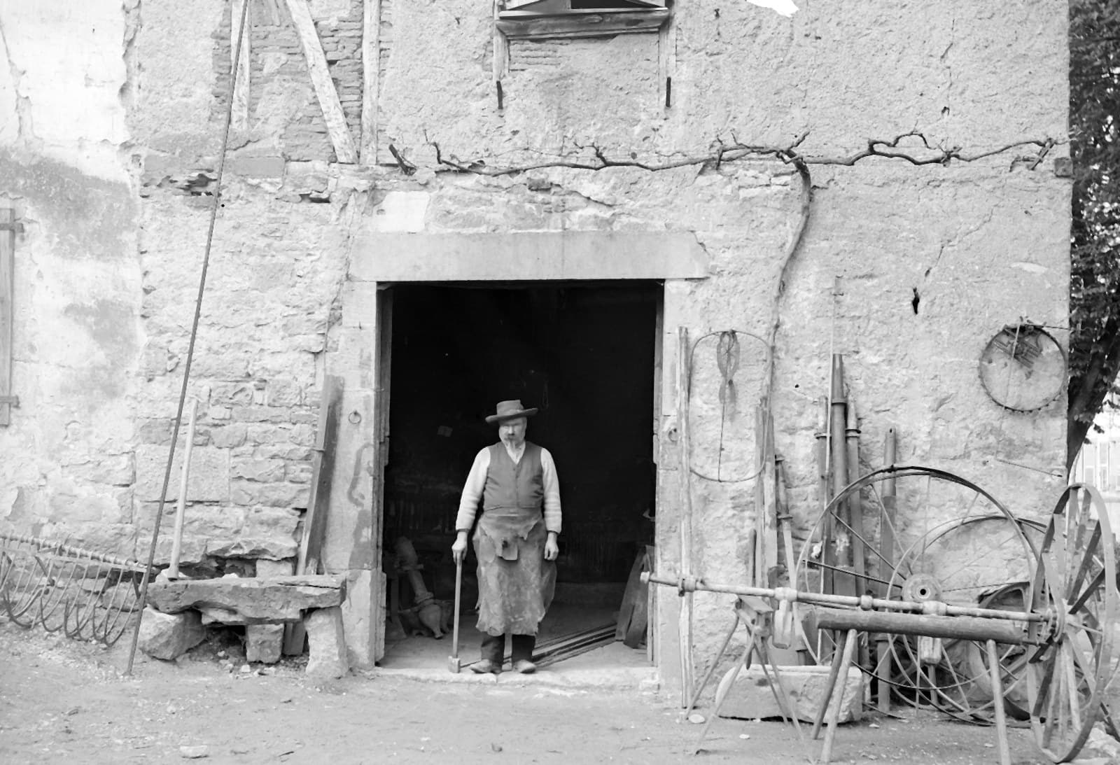 Un forgeron en tablier debout sur le seuil de sa forge, des roues et des outils appuyés contre le mur de pierre.