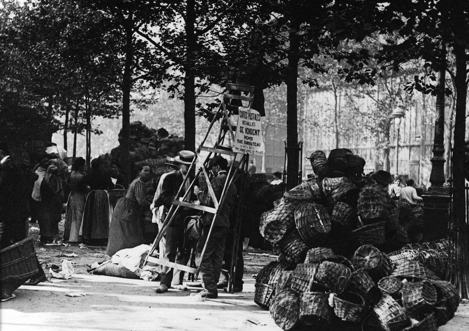 Aux Halles, le haut échafaudage de bois d’un photographe ambulant, coiffé de son appareil, avec une pancarte qui vend ses cartes postales, entre les passants et les piles de paniers.