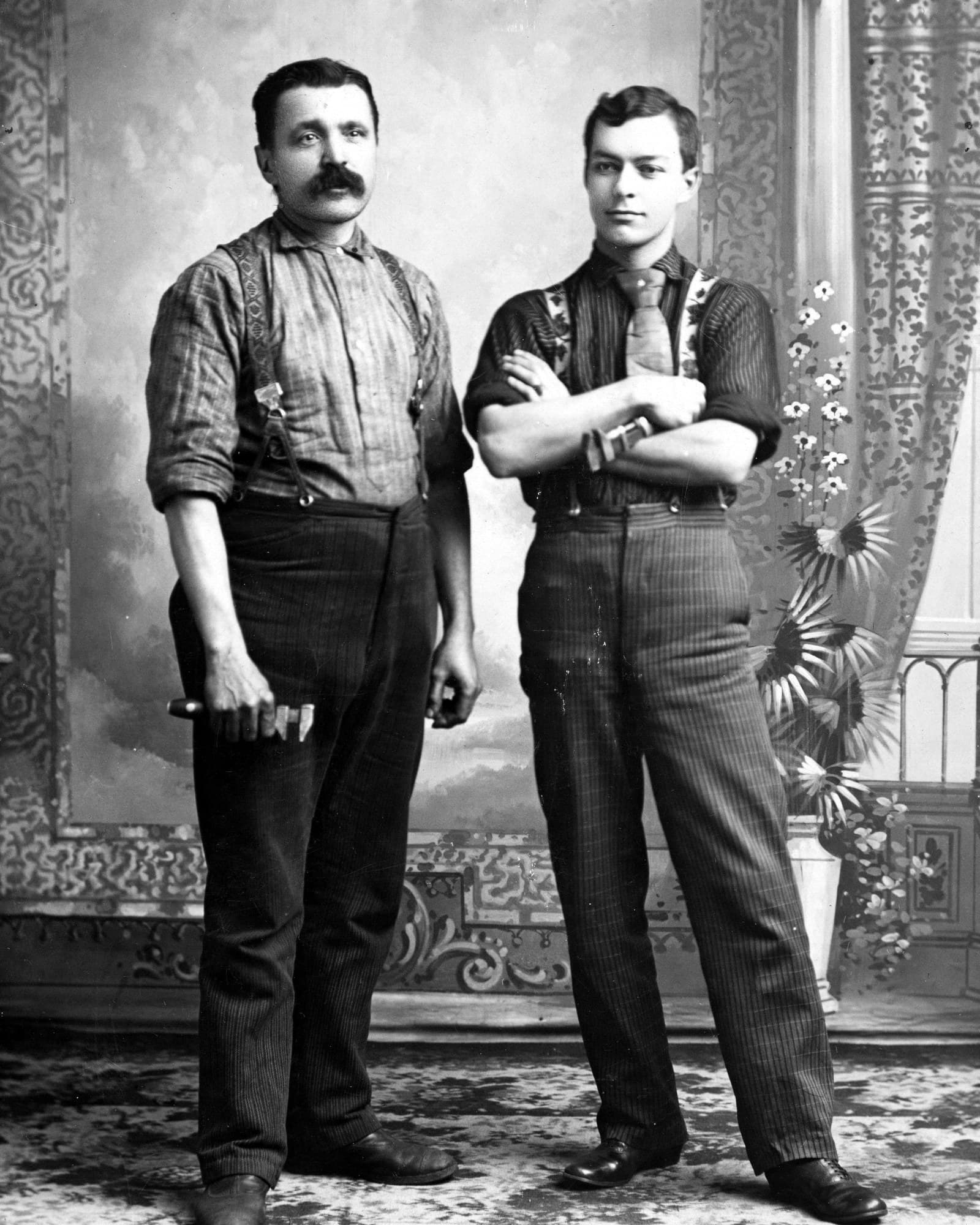 Portrait de deux plombiers en studio, en bretelles et en salopette, bras croisés, devant un décor peint.